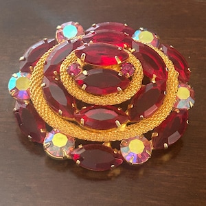 May include: A vintage brooch in a floral design. The brooch features a gold-tone metal base with a woven texture, and is adorned with oval and round red rhinestones, and iridescent accent stones.