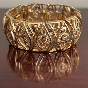 May include: A gold-toned bracelet with an openwork design. The bracelet features repeating teardrop-shaped links with an ornate, scroll-like pattern in the center of each. The bracelet is resting on a reflective, dark brown surface.