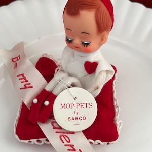 May include: Vintage Valentine's Day doll, a Mop-Pet by Sarco, Japan. The doll has a red cap and a white outfit with a red heart. It sits in a red felt bed with a white ribbon that says "Be my Valentine".