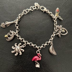 May include: Silver charm bracelet featuring various charms, including a flamingo, sun, alligator, palm tree, and a slice of citrus. The flamingo charm is pink with a small silver purse. The sun charm has the words "The Sunshine State".