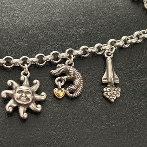 May include: Silver charm bracelet featuring a sun with a smiling face, a crocodile with a gold heart, and a rocket ship charm. The bracelet has a chain link design.
