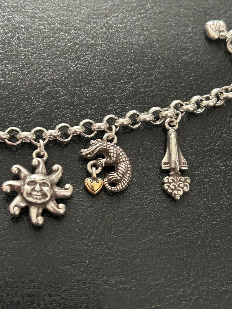 May include: Silver charm bracelet featuring a sun with a smiling face, a crocodile with a gold heart, and a rocket ship charm. The bracelet has a chain link design.