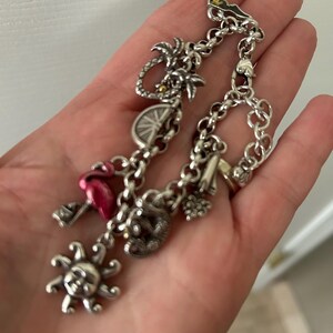 May include: Silver charm bracelet featuring various charms, including a sun, flamingo, palm tree, and a boot. The bracelet is made of linked chains and has a lobster clasp. The charms are detailed and add a whimsical touch.