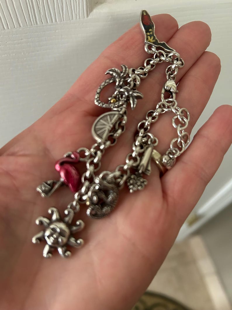May include: Silver charm bracelet featuring various charms, including a sun, flamingo, palm tree, and a boot. The bracelet is made of linked chains and has a lobster clasp. The charms are detailed and add a whimsical touch.