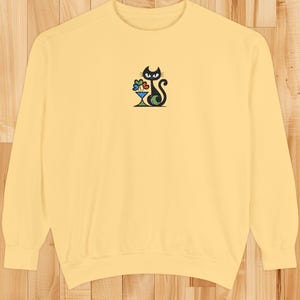 May include: A light yellow sweatshirt featuring an embroidered black cat design. The cat is depicted with a cocktail glass filled with colorful flowers. The design is centered on the chest of the long-sleeved crew neck top.