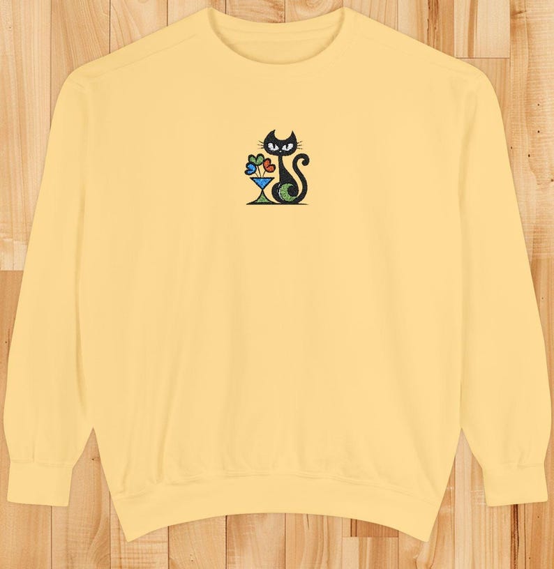 May include: A light yellow sweatshirt featuring an embroidered black cat design. The cat is depicted with a cocktail glass filled with colorful flowers. The design is centered on the chest of the long-sleeved crew neck top.