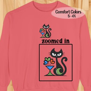 May include: A coral-colored sweatshirt with a black cat graphic holding flowers in a blue vase. The text "zoomed in" is below the cat graphic. The top right corner has the text "Comfort Colors S-4X".