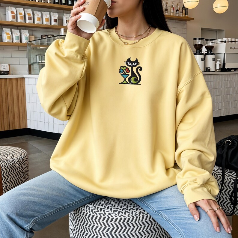 May include: A light yellow sweatshirt with a black cat design holding flowers in a vase. The cat is embroidered on the front. The sweatshirt has long sleeves and a crew neck.
