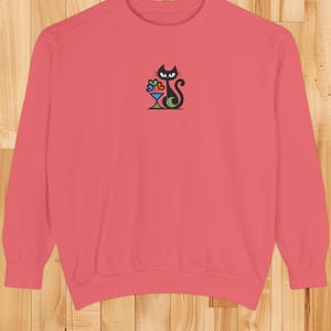 May include: A coral-colored sweatshirt featuring a black cat design with a cocktail glass. The cat has a curved tail and is holding a colorful cocktail. The design is embroidered on the front of the sweatshirt.