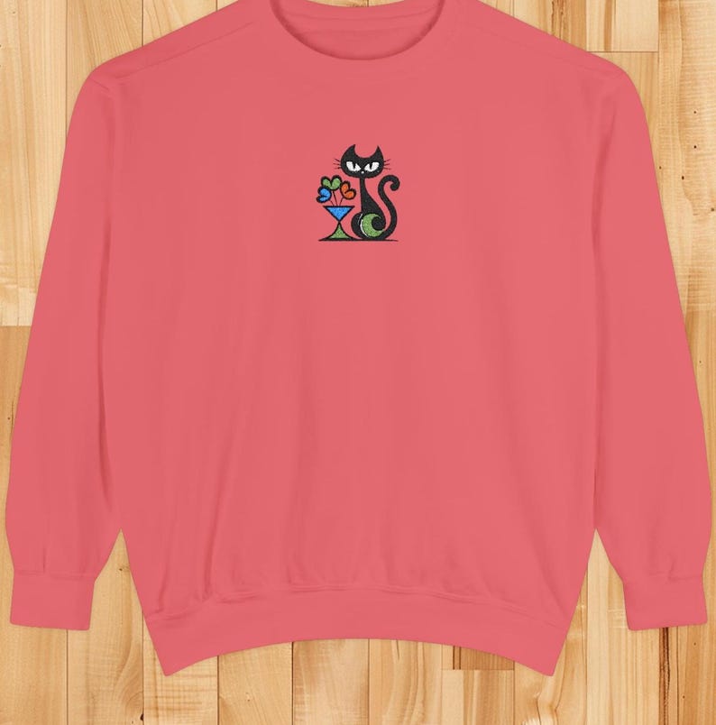May include: A coral-colored sweatshirt featuring a black cat design with a cocktail glass. The cat has a curved tail and is holding a colorful cocktail. The design is embroidered on the front of the sweatshirt.