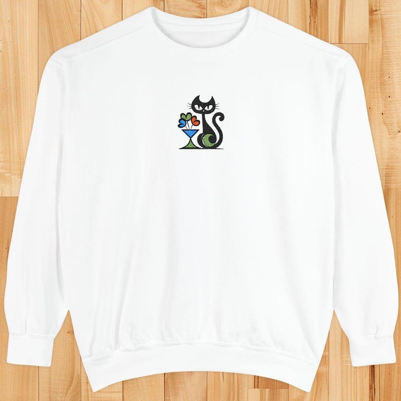 May include: White sweatshirt featuring a whimsical black cat design with a green, black, and white body, and a colorful flower arrangement. The cat is sitting in a martini glass. The sweatshirt has long sleeves and a crew neck.