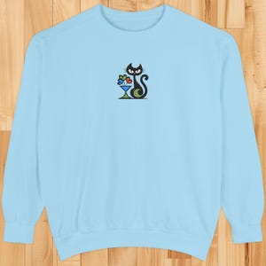 May include: Light blue sweatshirt with a black cat design holding flowers in a vase. The cat has a curved tail and is centered on the front of the long-sleeved top. The flowers are in shades of blue, red, and yellow.