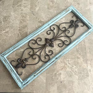 May include: Rectangular wall decor with a distressed light blue frame. The center features a dark brown metal design with a fleur-de-lis and floral accents. The frame is approximately 24 inches long and 12 inches wide.