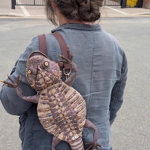 May include: A backpack shaped like a bearded dragon lizard, covered in intricate purple, gold, and brown beadwork. The lizard features yellow eyes, textured scales, and four legs extending outward. It includes brown fabric straps and a zipper closure.