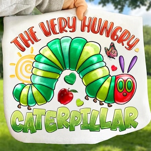 以下が含まれることがあります： 「THE VERY HUNGRY CATERPILLAR」の文字と、緑と赤のカラフルなイモムシのイラストが描かれた白いトートバッグ。デザインには赤いリンゴ、蝶、黄色い太陽が含まれています。