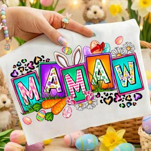 Op de afbeelding: Wit t-shirt met het woord "MAMAW" in kleurrijke blokletters, versierd met paaseieren, konijnenoren, wortels en pootafdrukken. Het t-shirt heeft een feestelijk, lentethema.