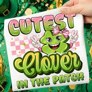 Może przedstawiać: Biała koszulka z motywem na Dzień Świętego Patryka. Koszulka zawiera napis "Cutest Clover in the Patch" z uśmiechniętą zieloną grafiką koniczyny w różowej kokardce i znakiem pokoju.