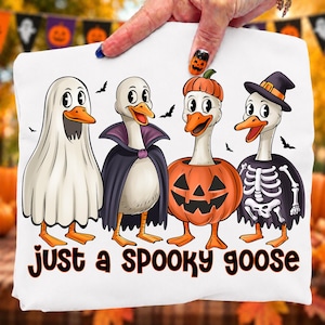 May include: A white t-shirt featuring a graphic of four geese in Halloween costumes: one as a ghost, one as a vampire, one inside a pumpkin, and one as a skeleton wearing a witch hat. The text below reads "just a SPOOKY goose" in black.