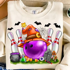 May include: Cream-colored sweatshirt featuring a Halloween-themed bowling design. A purple bowling ball wears an orange witch hat. The design includes bowling pins, bats, a spider web, and other Halloween elements.