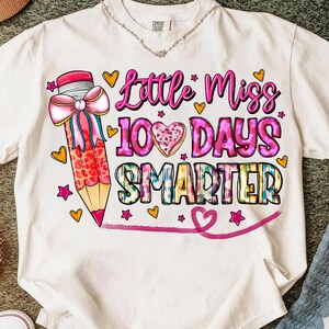 Op de afbeelding: Wit T-shirt met een kleurrijke afbeelding. Het ontwerp toont een grote potlood met een roze strik, de woorden "Little Miss 100 Days Smarter", harten en sterren. Het shirt is gemaakt van een zacht materiaal.