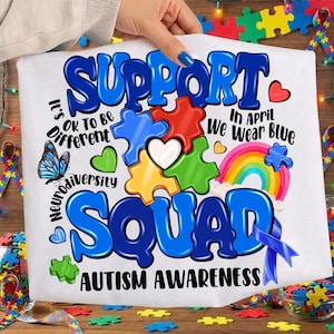 Op de afbeelding: Wit t-shirt met een kleurrijke afbeelding ter ondersteuning van autismebewustzijn. Het ontwerp bevat puzzelstukjes, een regenboog, een vlinder en de woorden "Support Squad" en "Autism Awareness". De zin "In April We Wear Blue" is ook te zien.