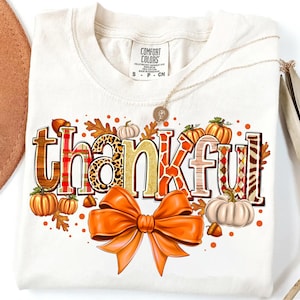 May include: Cream-colored t-shirt with the word "thankful" in a fall-themed design. The word is decorated with pumpkins, leaves, and an orange bow. A gold necklace with a pendant is also visible.
