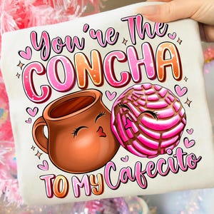 May include: A white t-shirt with a colorful graphic featuring a brown mug and a pink concha pastry, with the text "You're The Concha To My Cafecito" in pink and orange. The design includes hearts and stars.