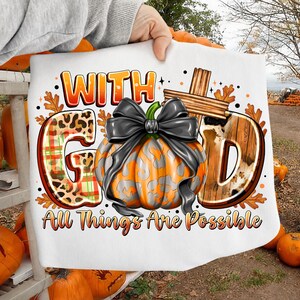 May include: A white t-shirt featuring a graphic with the text "WITH GOD" and "All Things Are Possible". The design includes a central orange pumpkin with a black bow, a wooden cross, and letters filled with leopard print, plaid, and cow patterns.