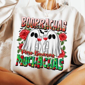 May include: White sweatshirt with a graphic design featuring two ghost figures holding wine glasses. The text "Borrachas Pero Buenas Muchachas" is displayed in a colorful, floral font, with red roses and green leaves.