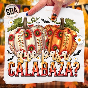 May include: A white t-shirt featuring two large pumpkins with intricate floral patterns in red, orange, and green. The text "que pasa, CALABAZA?" is printed in a bold, gradient font below the pumpkins, surrounded by small white daisies and black bats.