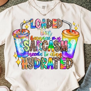 Może przedstawiać: Biała koszulka z kolorowym wzorem graficznym. Wzór przedstawia dwa kubki z napojami ze słomkami oraz napis "LOADED Tea because my SARCASM needs to stay HYDRATED". Tekst i kubki mają różne wzory i kolory.