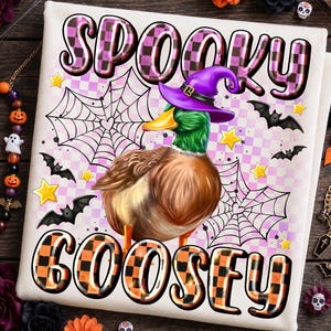 May include: A white fabric item featuring a mallard duck wearing a purple witch hat. The text SPOOKY GOOSEY is printed in a checkered pattern. The design includes black spiderwebs, small black bats, and yellow stars on a light purple checkered background.