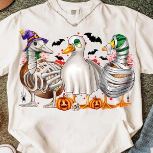 May include: A cream-colored short-sleeve t-shirt featuring a graphic of three ducks dressed for Halloween. One duck wears a purple witch hat and skeleton bones, one is a ghost, and one is a mummy. Two jack-o'-lanterns, a skull, and bats are included.