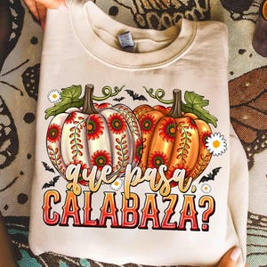 May include: A beige crewneck sweatshirt featuring a graphic of two pumpkins decorated with red and green floral patterns. The design includes small white flowers, black bats, and the text "que pasa CALABAZA?" in a stylized orange and red font.