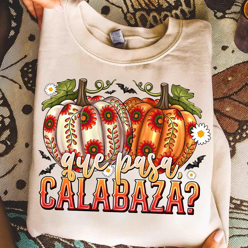 May include: A beige crewneck sweatshirt featuring a graphic of two pumpkins decorated with red and green floral patterns. The design includes small white flowers, black bats, and the text "que pasa CALABAZA?" in a stylized orange and red font.