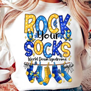 May include: White sweatshirt with the text "Rock Your Socks" in blue and yellow letters with a blue and yellow ribbon with a puzzle piece. The text "World Down Syndrome" is below the main text. There are four socks hanging on a clothesline with a blue and yellow ribbon on each sock.