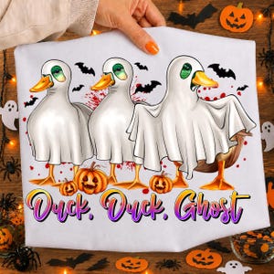May include: A white t-shirt featuring a graphic of three ducks wearing white ghost costumes. The ducks are positioned above two orange jack-o'-lantern pumpkins and black bats. The text "Duck, Duck, Ghost" is printed in a purple and orange gradient.