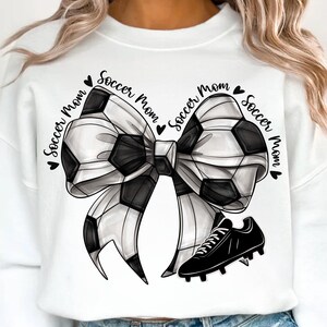 May include: White sweatshirt with a black and white soccer ball bow design and a black soccer cleat. The words "Soccer Mom" are written around the bow in a circular pattern. A cute and sporty design.