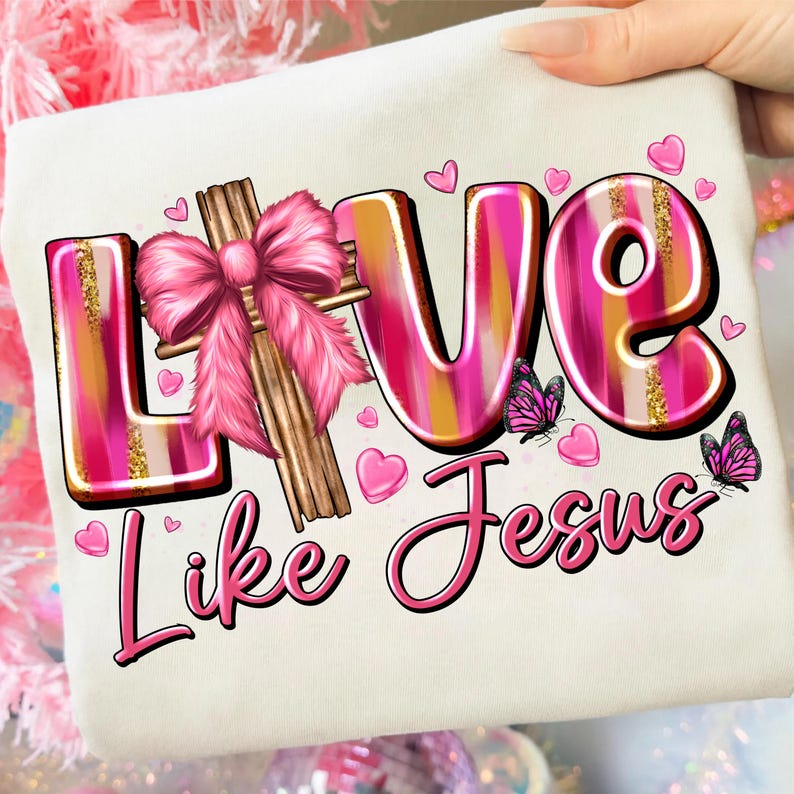May include: Cream-colored t-shirt with the words "LOVE Like Jesus" in pink and gold. The word "LOVE" features a pink bow and cross design. Pink hearts and butterflies are scattered around the text.