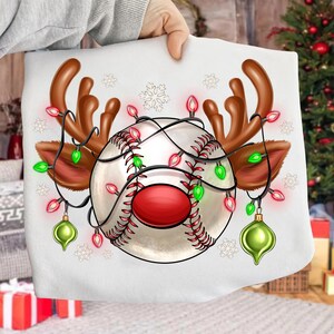 Pode incluir: Camisola branca com um basebol decorado com chifres de rena e luzes de Natal. O basebol tem um centro vermelho e é adornado com luzes e enfeites vermelhos e verdes. Chifres e orelhas castanhos em ambos os lados.