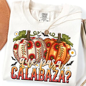 May include: A cream-colored t-shirt featuring a graphic of two pumpkins decorated with red and green floral patterns. The text "que pasa CALABAZA?" is printed below the pumpkins in a stylized font. Small bats and white daisies surround the pumpkins.