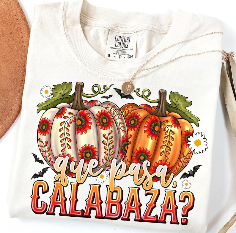 May include: A cream-colored t-shirt featuring a graphic of two pumpkins decorated with red and green floral patterns. The text "que pasa CALABAZA?" is printed below the pumpkins in a stylized font. Small bats and white daisies surround the pumpkins.