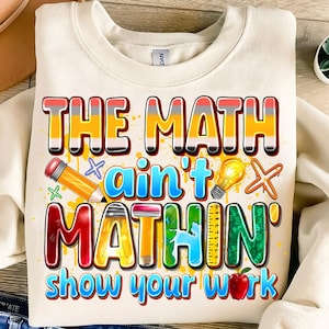 Puede incluir: Sudadera color crema con un gráfico colorido que dice "THE MATH ain't MATHIN' show your work". El diseño incluye un lápiz, una bombilla, una manzana y una regla.