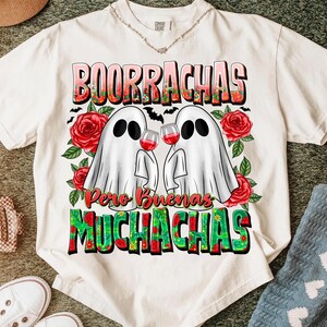 May include: White t-shirt featuring a graphic design with the words "BOORRACHAS" and "MUCHACHAS" in colorful letters. Two ghost figures are holding wine glasses, surrounded by red roses and bats.
