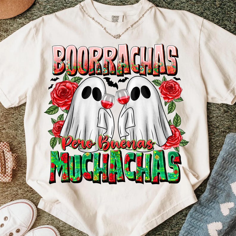 May include: White t-shirt featuring a graphic design with the words "BOORRACHAS" and "MUCHACHAS" in colorful letters. Two ghost figures are holding wine glasses, surrounded by red roses and bats.