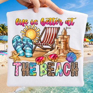 以下が含まれることがあります： 「life is better at THE BEACH」という文字が書かれたカラフルなグラフィックがプリントされた白いTシャツ。デザインには、黄色い太陽、赤と白のストライプのビーチチェア、砂の城、青いビーチサンダル、ヒトデ、サングラス、貝殻が含まれています。