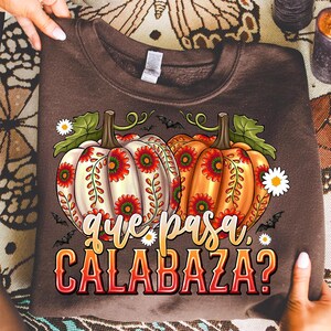 May include: A dark brown crewneck sweatshirt featuring a graphic of two pumpkins, one white and one orange, decorated with red and green floral patterns. The text "que pasa, CALABAZA?" is printed in a stylized font below the pumpkins with small bats.