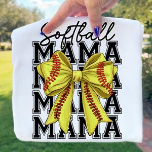 May include: White sweatshirt with the words "Softball Mama" in black script and block letters. A large yellow bow with red stitching, resembling a softball, is centered on the design. The sweatshirt is held up by a hand.