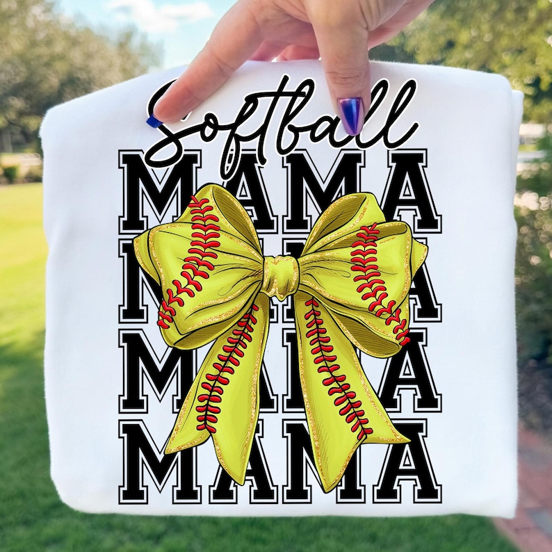 May include: White sweatshirt with the words "Softball Mama" in black script and block letters. A large yellow bow with red stitching, resembling a softball, is centered on the design. The sweatshirt is held up by a hand.