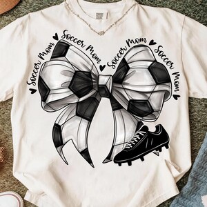 May include: White t-shirt featuring a soccer ball bow design with the words "Soccer Mom" and a black soccer cleat. The bow is black and white, and the text is in a script font.
